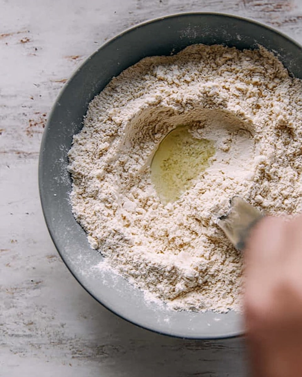 A close-up view of a gray bowl filled mostly with light beige flour, with a small pool of clear liquid in the center, and a woman's hand partially visible on the right side, about to mix the ingredients, set on a white marbled surface photo taken with an iphone --ar 4:5 --v 7