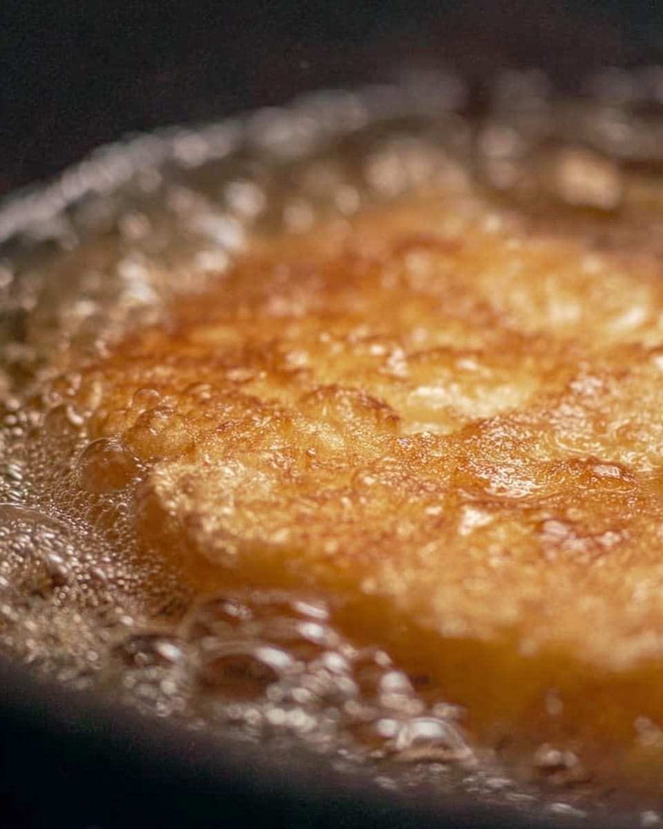 The image shows a close-up of a golden brown piece of food frying in bubbling hot oil inside a dark pan. The food has a slightly rough surface with a crispy texture visible on top. The oil around it is foaming and moving quickly, giving a sense of heat and cooking in progress. The pan edges are barely visible, focusing mainly on the frying food and the bubbles of oil surrounding it. photo taken with an iphone --ar 4:5 --v 7