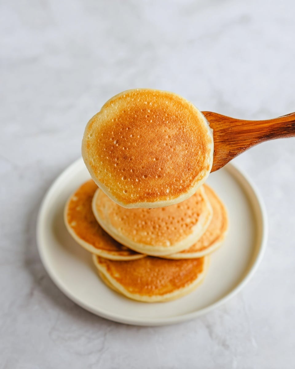 The image shows a stack of four golden brown pancakes on a plain white plate with three pancakes lying flat and one pancake being held over the plate by a wooden spatula. Each pancake has a smooth texture with a slightly crispy edge and small bubbles on the surface, giving it a light and fluffy look. The background is a white marbled texture that contrasts softly with the warm colors of the pancakes. Photo taken with an iphone --ar 4:5 --v 7
