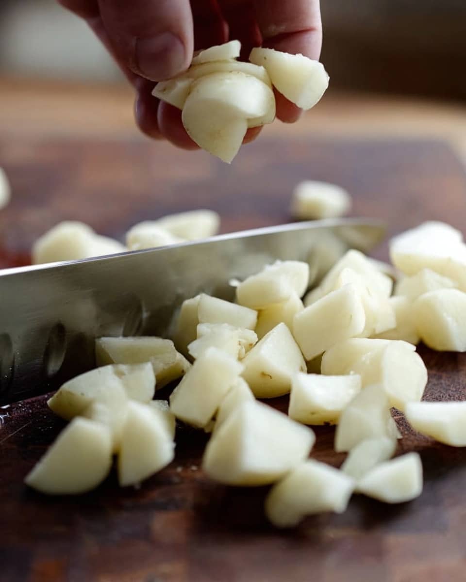 A close-up view showing a woman's hand holding a small pile of peeled potato pieces above a dark wooden cutting board. The potato pieces are light and creamy white, with smooth edges, and are being moved by a shiny, large kitchen knife with a thin blade positioned horizontally. More chopped potato pieces are scattered on the cutting board below, creating a layered effect of irregular, small white chunks over the dark wood grain. The background is slightly blurred to keep the focus on the knife and potatoes. Photo taken with an iphone --ar 4:5 --v 7