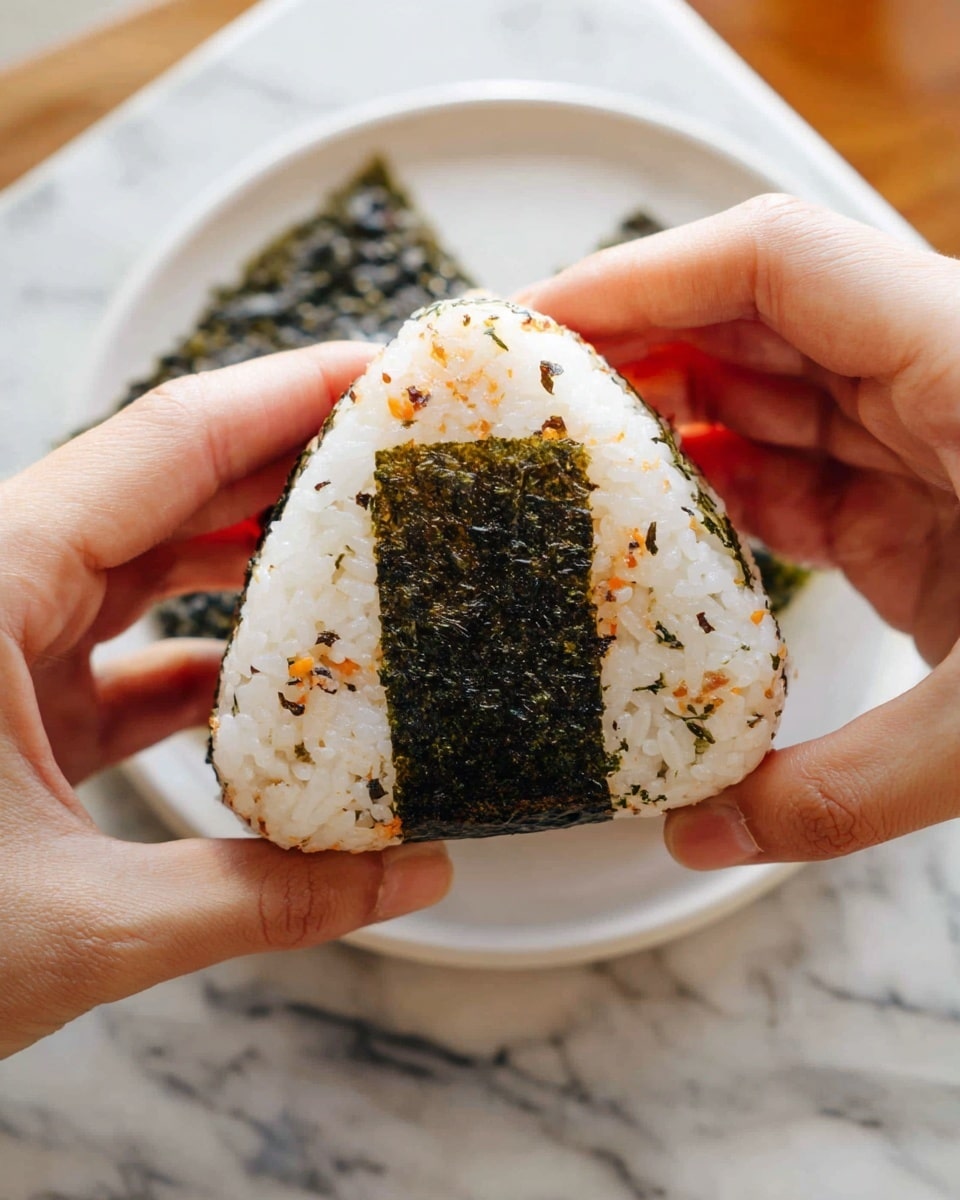 A triangular rice ball held by two woman's hands, with a white sticky rice outer layer sprinkled with small orange, black, and green bits around the edges, and a square piece of dark green seaweed wrapped around the middle front. The background shows a white plate with a sheet of seaweed on a white marbled surface. Photo taken with an iphone --ar 4:5 --v 7
