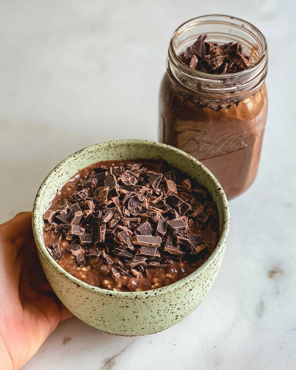 A light green speckled bowl filled with a two-layered dish stands on a hand against a white marbled surface. The bottom layer is a rich, thick dark brown mixture with bits of lighter brown pieces mixed throughout. On top, there is a generous layer of unevenly chopped dark chocolate pieces in various sizes and shapes, spreading over the surface. Behind the bowl, there is a mason jar filled with the same dark brown mixture, capped with a silver lid. The overall scene has a clean and simple look with a focus on the bowl's rich chocolatey content photo taken with an iphone --ar 4:5 --v 7