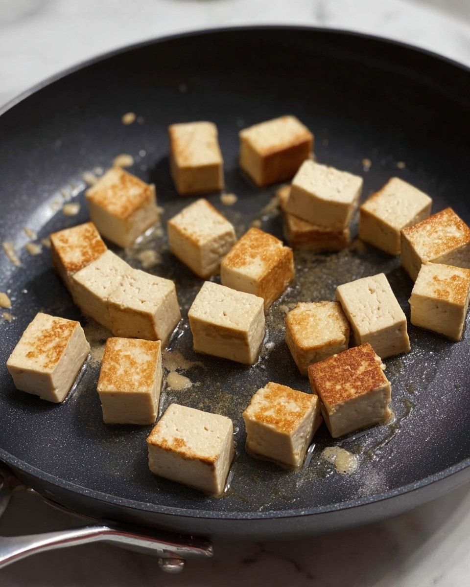 The image shows a cooking pan with 18 small cubes of tofu inside. Each tofu cube is light beige with a golden brown crust on some sides, showing they are frying. The cubes have a smooth but slightly textured surface and are scattered in the non-stick pan with some oil droplets visible. The pan is shown close up against a white marbled background. photo taken with an iphone --ar 4:5 --v 7