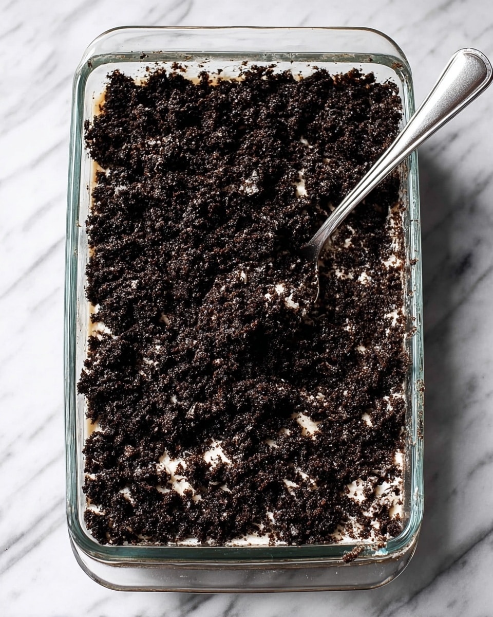 A clear glass rectangular dish filled with a dark crumbly mixture that has small patches of white creamy bits spread throughout. The mixture looks rough and uneven in texture, pressed down to form one thick layer covering the bottom of the dish. A silver spoon rests on top of the mixture near the bottom right corner, with some of the mixture sticking to it. The dish is placed on a white marbled surface. photo taken with an iphone --ar 4:5 --v 7