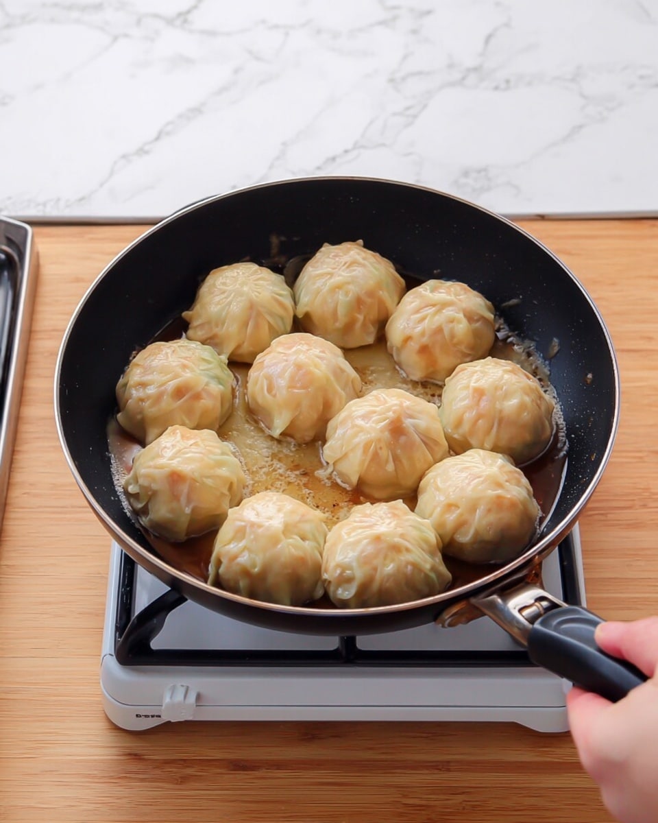 A black frying pan is filled with nine round dumplings arranged in a 3 by 3 pattern. Each dumpling has a light tan color with a slightly translucent skin and a soft, smooth texture, showing some folds near the top where the dough is pinched. The dumplings are sitting in a thin, light brown sauce that glistens under the light. A woman's hand is holding the black pan handle on the right side, and the pan is on a white portable gas stove. The stove and the wooden surface beneath it contrast with the white marbled texture background. Photo taken with an iphone --ar 4:5 --v 7