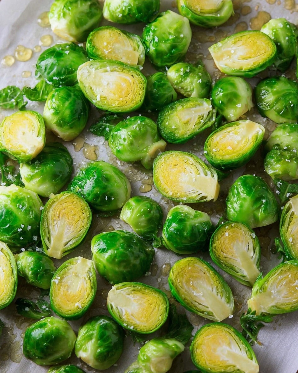 The image shows many halved Brussels sprouts spread out on baking paper over a white marbled surface. The Brussels sprouts have a bright green color with a slight yellow tint in the middle layers. Some loose outer leaves are scattered around the halved pieces. Tiny clear droplets of oil and small grains of salt are visible on the Brussels sprouts and paper, adding a shiny texture and contrast to the matte green leaves. photo taken with an iphone --ar 4:5 --v 7