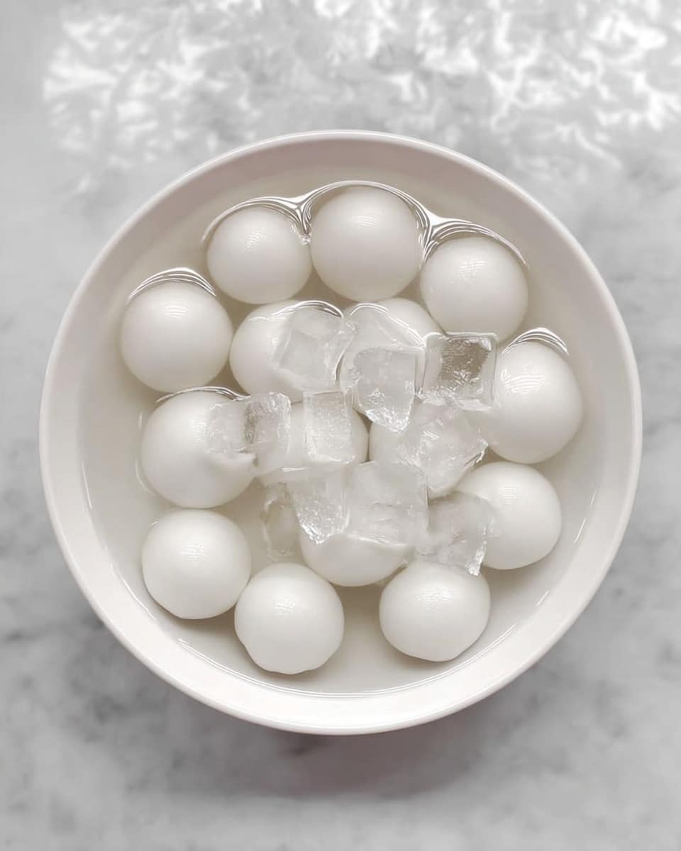 A white round bowl filled with about sixteen smooth, white, round dumplings submerged in water, with clear ice cubes floating among them. The water gives a glossy, wet look to the dumplings and ice. The bowl sits on a white marbled surface. photo taken with an iphone --ar 4:5 --v 7