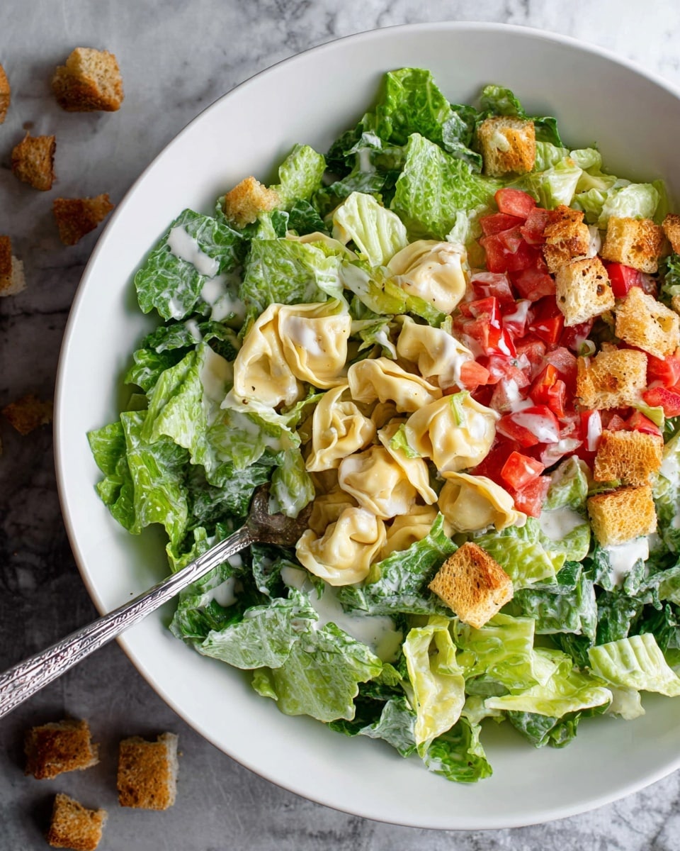 A white bowl holds a fresh salad with three main layers: the bottom layer is bright green chopped Romaine lettuce, the middle layer has folded pale yellow tortellini pasta, and the top layer includes small red tomato cubes and crunchy golden brown croutons scattered around. A creamy white dressing is drizzled lightly over the salad, giving a shiny look to the lettuce and pasta. A silver fork rests inside the bowl, partially buried in the salad, all against a white marbled textured surface, with some croutons scattered outside the bowl. photo taken with an iphone --ar 4:5 --v 7