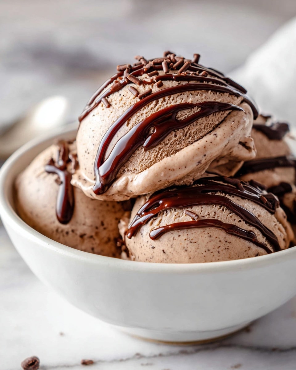 The image shows a close-up of three scoops of smooth, creamy chocolate ice cream stacked slightly on each other in a white bowl. The ice cream is light brown with tiny chocolate bits mixed inside, making the texture look rich and soft. Thin streams of dark chocolate sauce drizzle over the top scoop, adding a glossy dark contrast that zigzags down the sides. Small chocolate sprinkles are scattered on and around the ice cream, giving extra texture and a playful look. The background is a white marbled surface with soft lighting highlighting the shiny chocolate sauce and creamy ice cream texture. photo taken with an iphone --ar 4:5 --v 7