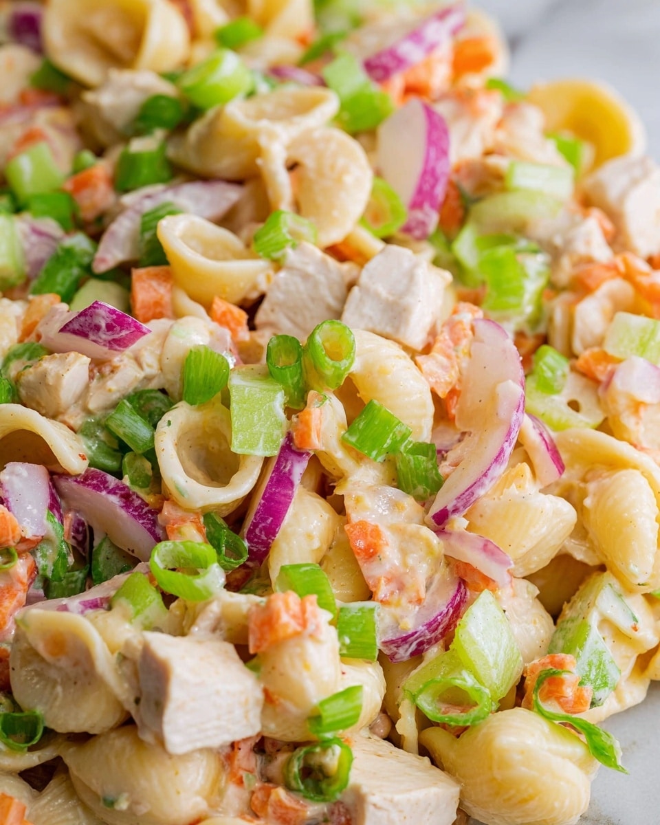 This close-up image shows a layered pasta salad with creamy dressing. The base layer is small shell pasta, light beige in color and smooth textured, mixed evenly with cubes of pale white chicken pieces. Small chunks of light green celery and orange carrots are scattered throughout, adding a crunchy texture. There are thin, round slices of purple-skinned radishes adding pops of color. The salad is topped with thinly sliced bright green scallions, providing a fresh touch. The whole dish is on a white marbled surface. photo taken with an iphone --ar 4:5 --v 7