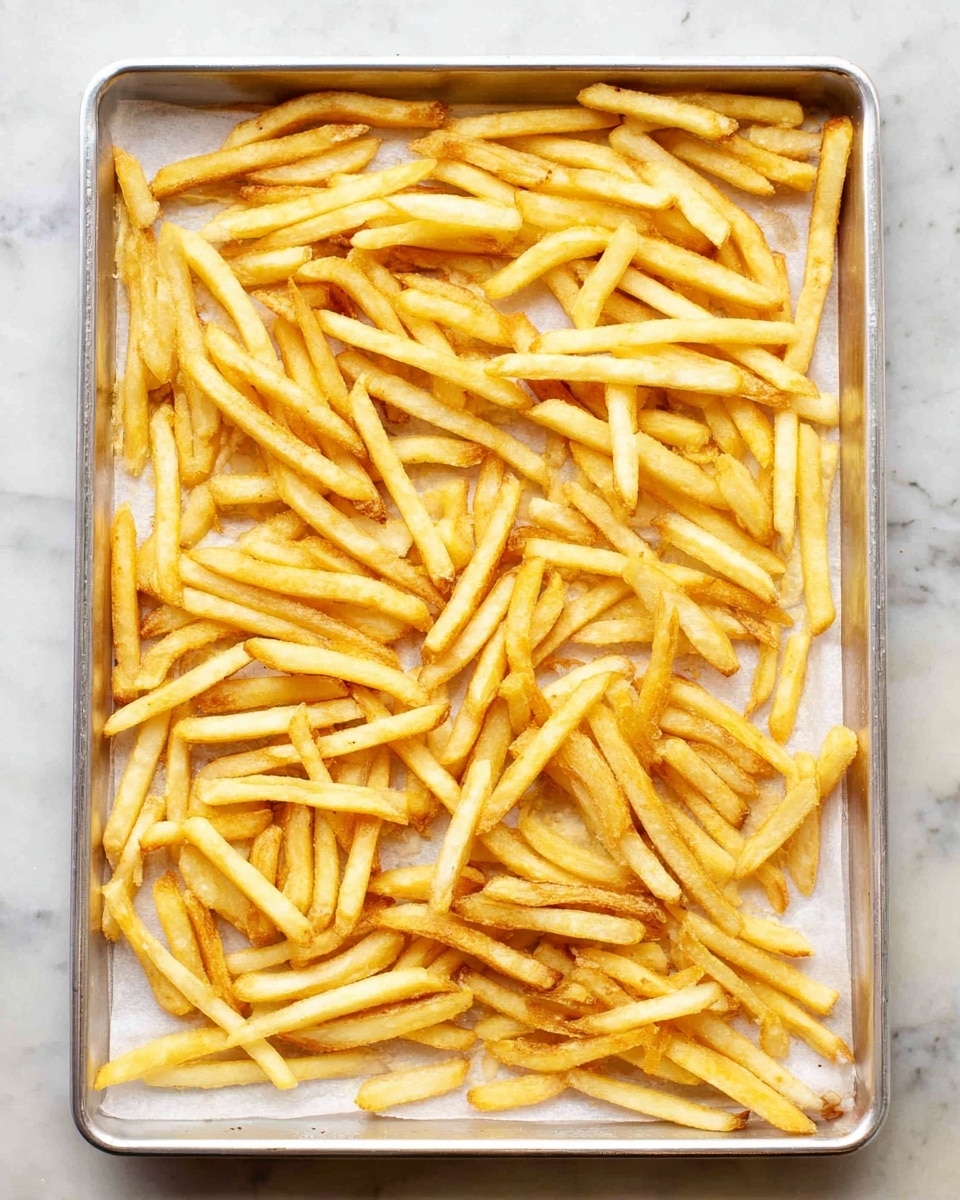A silver baking tray holds a single layer of golden-yellow French fries spread evenly across white parchment paper. The fries have a slightly crispy texture with some edges showing a light brown tint, indicating light crispiness. The background is a white marbled surface that contrasts softly with the warm colors of the fries. The photo is clear and bright, showing the details of each fry's thin and straight shape. Photo taken with an iphone --ar 4:5 --v 7