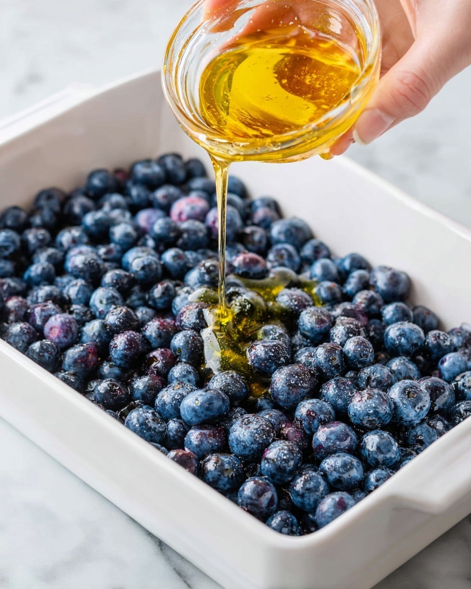 A white rectangular dish is filled with a thick layer of fresh blueberries with a mix of dark and light blue colors, some with a slight shine and powdery texture. Over the blueberries, a woman's hand is pouring golden honey from a small clear glass container, the honey catching light and adding a glossy liquid layer over the fruit. The dish is placed on a white marbled surface. photo taken with an iphone --ar 4:5 --v 7
