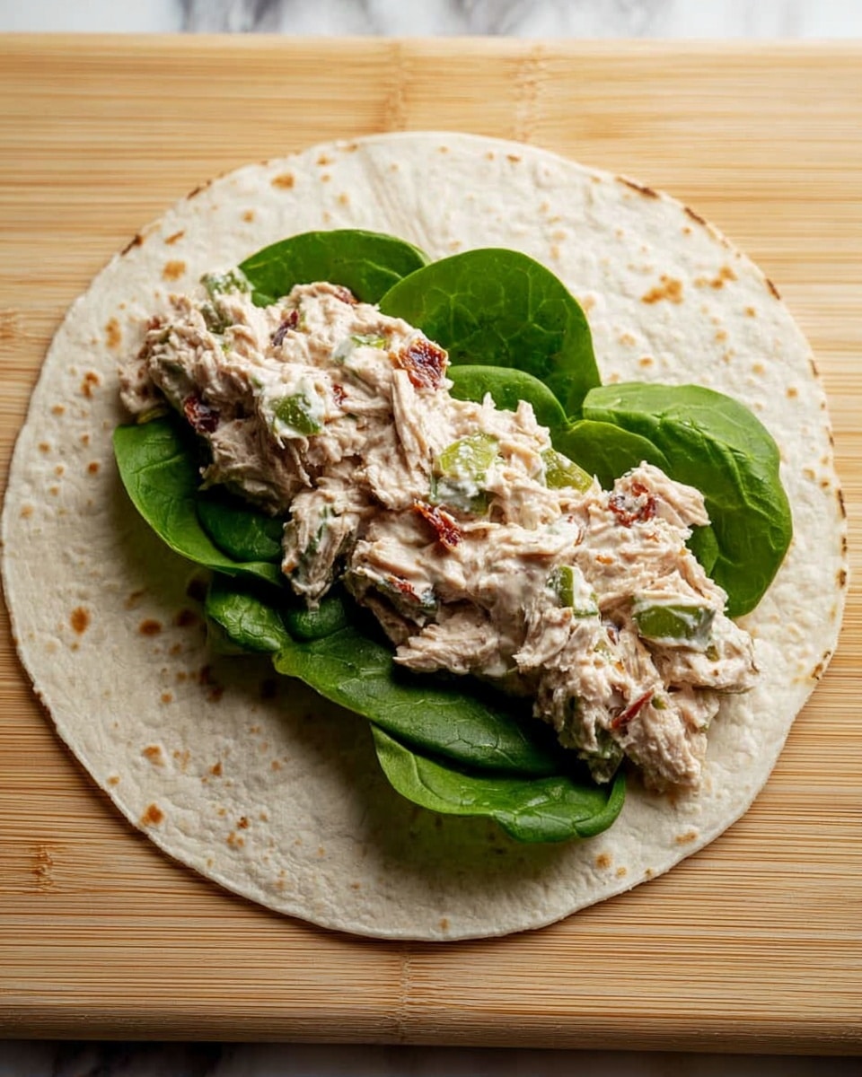 A single white tortilla wraps the base layer, lying flat with light brown toasted spots scattered evenly across its surface. On top of the tortilla are some fresh, dark green spinach leaves, arranged in a single layer. Over the spinach, there is a thick, creamy mixture of tuna salad with visible small pieces of celery and bits of sun-dried tomatoes, giving it a mix of light pinkish, green, and reddish-brown colors. The whole arrangement is placed on a light wooden board, with a white marbled texture softly visible in the background. photo taken with an iphone --ar 4:5 --v 7