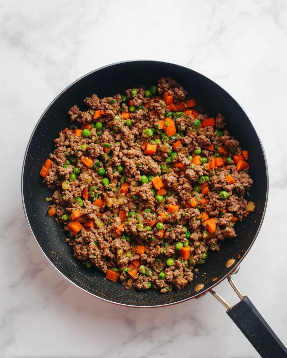 A black frying pan filled with a mix of cooked ground meat, bright orange carrot pieces, and small green peas evenly spread throughout. The meat is crumbly and browned, contrasting with the vibrant colors of the vegetables. The pan has a long black handle resting on a white marbled surface. The scene is lit naturally with a clean, simple background. photo taken with an iphone --ar 4:5 --v 7