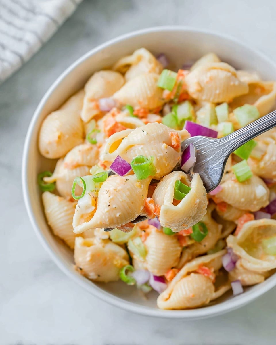 A close-up view of a bowl filled with creamy pasta salad made of large shell pasta that is light orange in color, mixed with small pieces of diced celery in pale green, finely chopped orange carrots, and thin slices of purple-red onion. Bright green scallion rings are scattered on top, adding freshness. The salad is coated in a light creamy dressing giving the pasta shells a smooth, slightly shiny texture. A silver fork is holding a few pasta shells layered with small vegetable pieces, positioned near the bowl’s edge. The bowl is white ceramic and set on a white marbled surface. Photo taken with an iphone --ar 4:5 --v 7