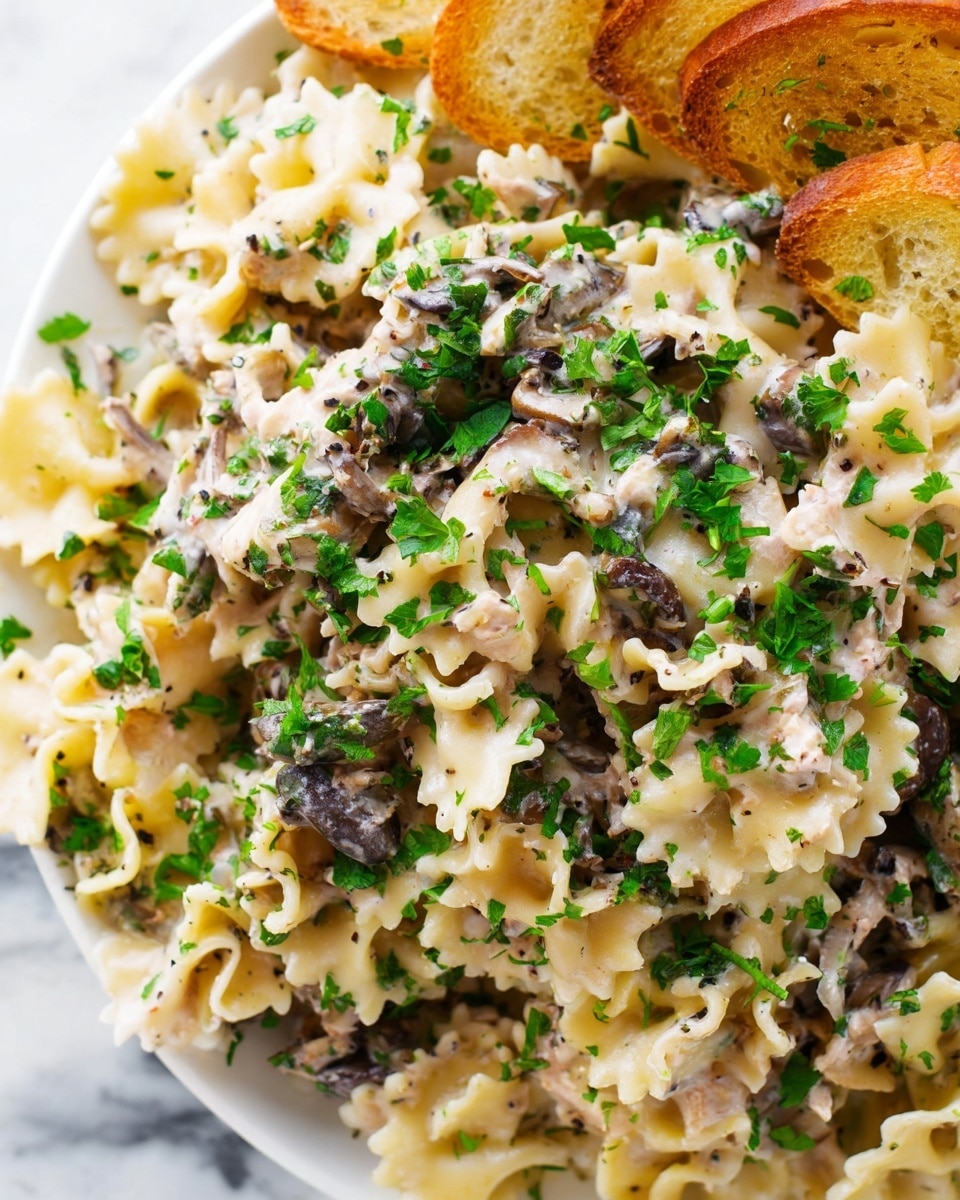 The image shows a close-up of a creamy pasta dish with ruffled edges. The pasta layers are mixed with small pieces of dark brown mushrooms and bits of chicken, all coated in a light creamy sauce. Bright green chopped herbs are sprinkled generously over the top, adding fresh color contrast. Some toasted, golden-brown bread slices are visible on the side of the white plate, resting on a white marbled surface. The textures of creamy sauce and soft pasta with bits of mushrooms and herbs create a rich and fresh look. photo taken with an iphone --ar 4:5 --v 7