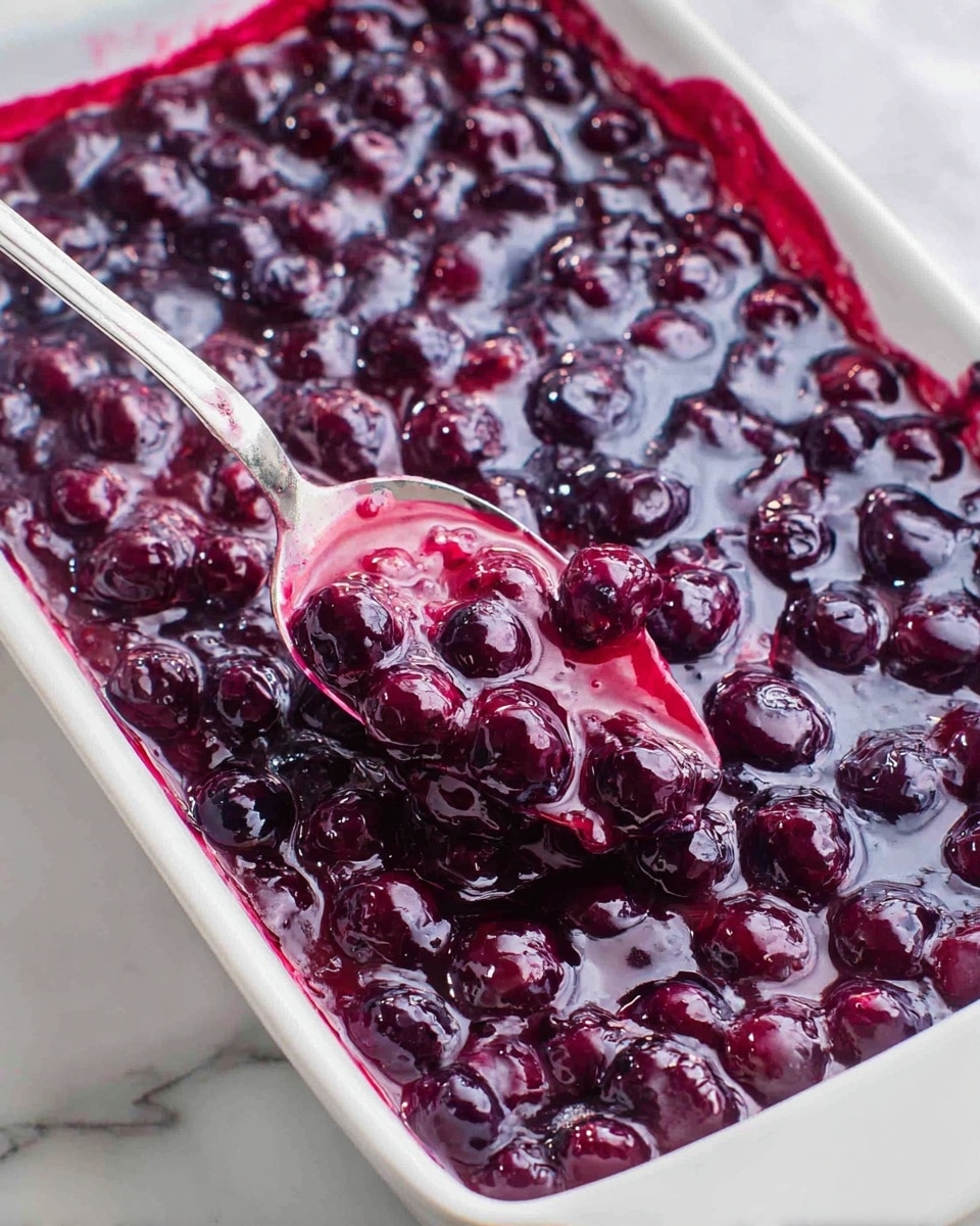 A close-up view of a white rectangular dish filled with a thick, glossy mixture of dark purple blueberries in a shiny sauce, creating a textured top layer with whole berries visible throughout. A silver spoon rests partially inside the dish, scooping some of the berry sauce, showing the rich purple jelly-like texture that clings to the spoon. The background features a white marbled surface beneath the dish. photo taken with an iphone --ar 4:5 --v 7