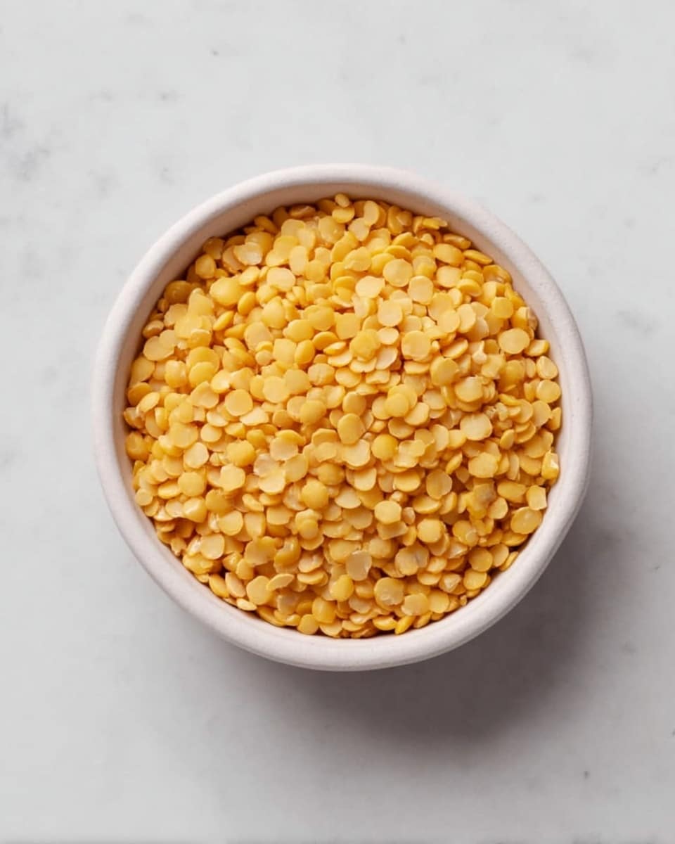 A small round white bowl filled with yellow split lentils, with a smooth and slightly shiny texture. The lentils are small, round, and evenly spread inside the bowl. The bowl is placed on a white marbled surface, photographed from above. photo taken with an iphone --ar 4:5 --v 7