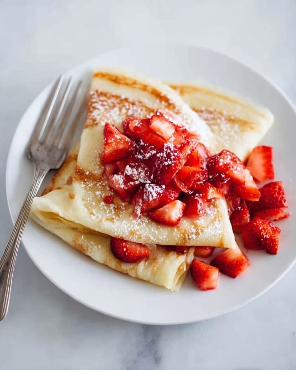 A white plate holds three folded crepes stacked in the center. The crepes are light golden brown with soft, smooth textures. On top of the crepes, there is a pile of chopped bright red strawberries, with a few more strawberry pieces scattered around the plate. A light dusting of white powdered sugar is sprinkled over the strawberries and crepes. The scene is set on a white marbled surface with a silver fork resting on the left side of the plate. Photo taken with an iphone --ar 4:5 --v 7