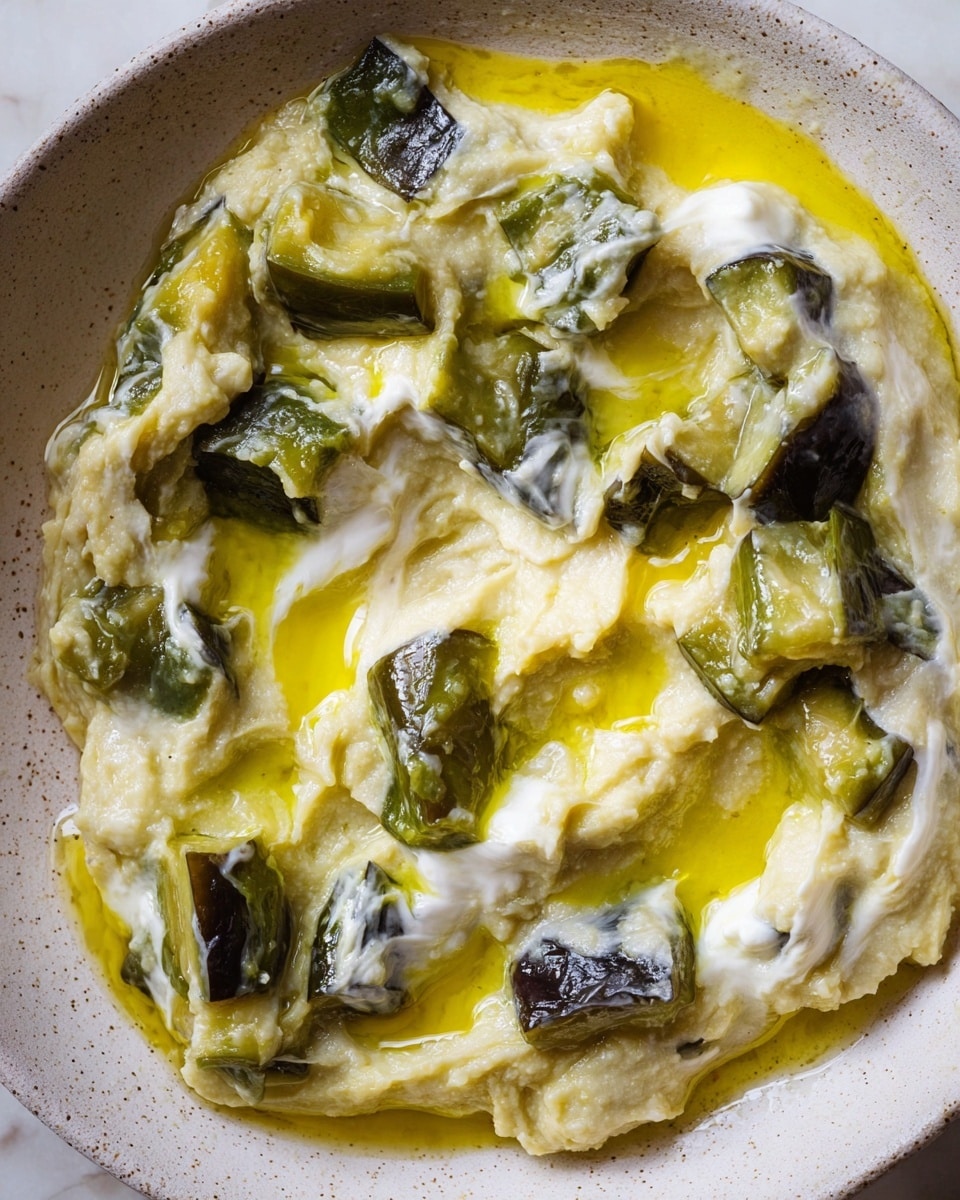The image shows a close-up of a creamy mixture with chunks of soft, cooked eggplant that have a dark green skin and pale, tender insides. The dish contains thick, pale yellow layers of mashed eggplant mixed with white yogurt swirls spread unevenly throughout. Shiny olive oil is drizzled over the top, creating bright yellow glossy patches that blend into the smooth textures. The mixture sits in a white bowl with a slightly speckled surface, and the background is a white marbled texture. photo taken with an iphone --ar 4:5 --v 7